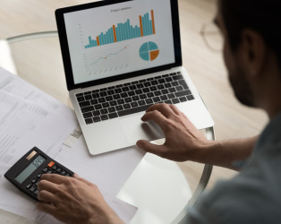 Imagem de um notebook com gráficos em tela, em cima de uma mesa de escritório com uma calculadora sendo utilizada por um homem, mostrado apenas de perfil e de costas que está trabalhando.
