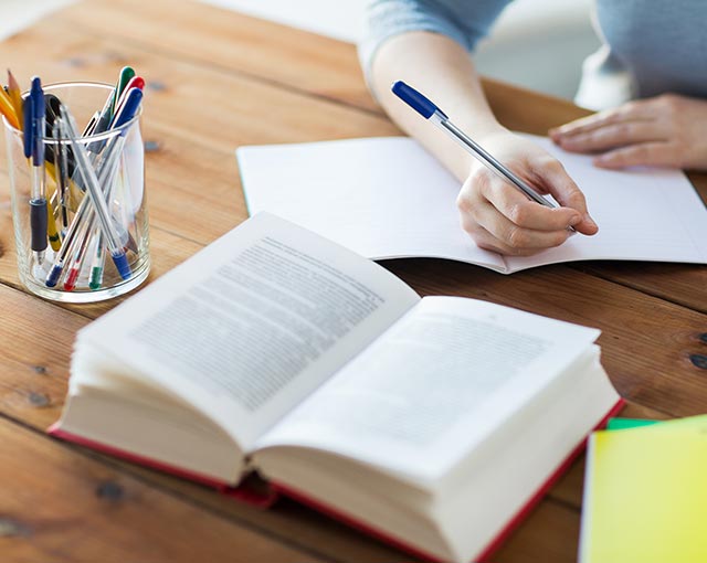 Publicações Imagem de uma pessoa escrevendo em um caderno em branco sob uma mesa de madeira com um livro aberto à sua frente e um porta canetas ao lado do livro cheio de canetas coloridas.